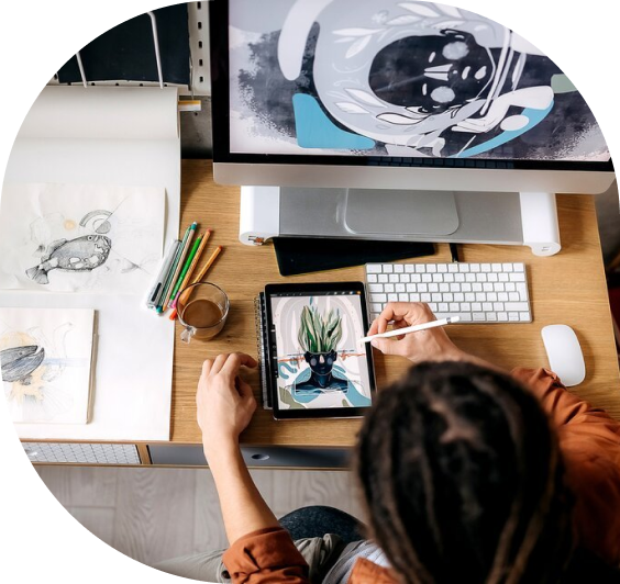 Overhead view of a digital artist using a stylus on a tablet to create character artwork, surrounded by sketches and a desktop monitor, representing creative design work.