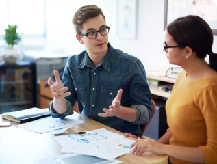 Young man and woman engaged in a professional conversation at a desk with data reports and a tablet, planning a project.