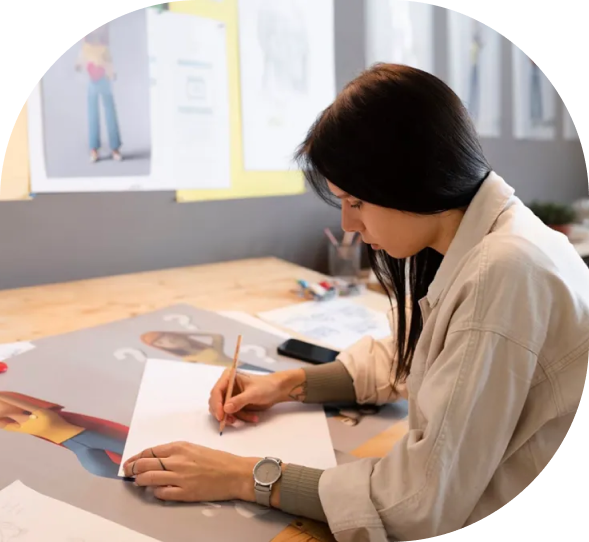 Female designer sketching character concepts on paper at a creative workspace, surrounded by fashion illustrations and reference materials pinned on the wall.