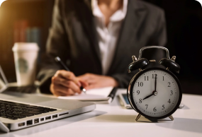 Classic black alarm clock on a desk beside a laptop and a person writing notes, symbolizing time management in a professional work environment.