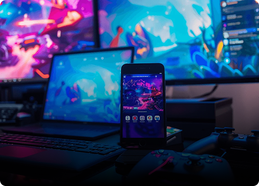 Colorful triple-monitor gaming setup with laptop, smartphone, and controllers showcasing a digital game interface.
